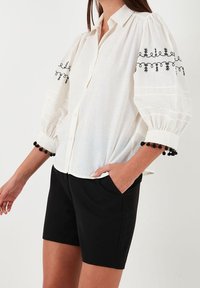 Chemise blanche légère avec des manches bouffantes, motif floral noir brodé sur les poignets et accents en pompons noirs. Associée à un short noir.