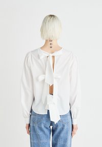 Blouse blanche à manches longues, dos ouvert, avec des nœuds noués. Portée avec un jean bleu orné de clous argentés dans un motif à carreaux.