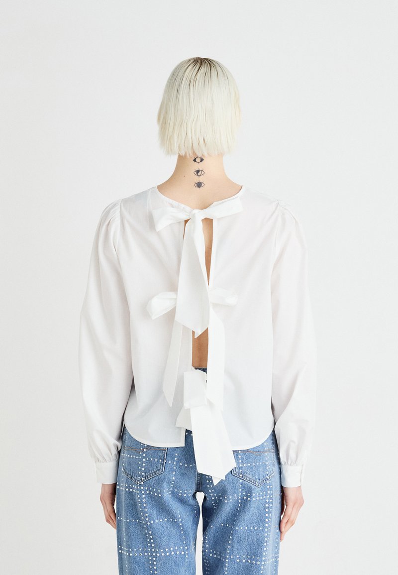 Blouse blanche à manches longues, dos ouvert, avec des nœuds noués. Portée avec un jean bleu orné de clous argentés dans un motif à carreaux.