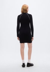 Μαύρο φόρεμα με ριμπ ύφασμα, μακριά μανίκια, υψηλός λαιμός και λευκή αντίθεση ραφών κατά μήκος των ραφών· εφαρμοστή σιλουέτα μέχρι το μέσο μηρού.