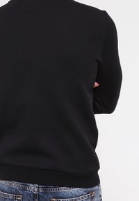 Sudadera negra de manga larga, con cuello redondo y puños acanalados. La tela parece suave y texturizada. Se lleva puesta sobre vaqueros azules desgastados.