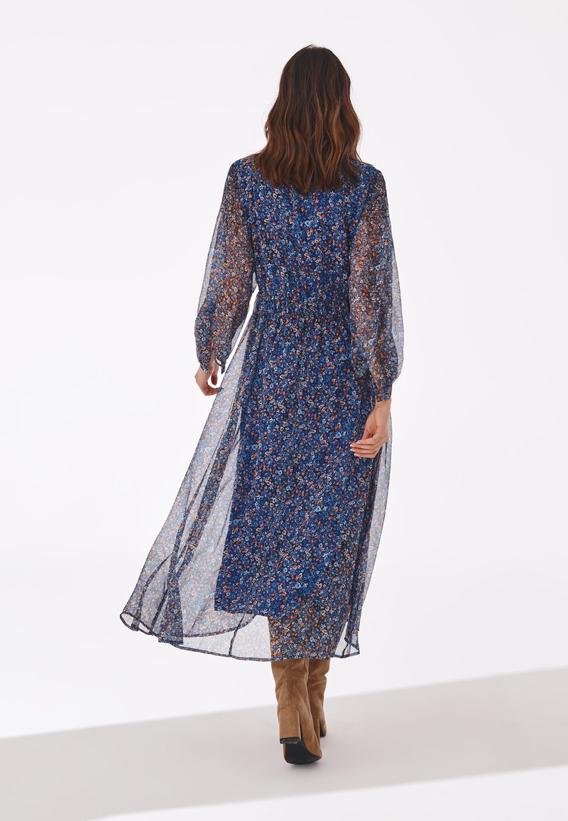 Robe longue et transparente à fleurs bleues avec de longues manches bouffantes et une taille élastique. Associée à des bottes en cuir suédé beige montant jusqu'au genou.