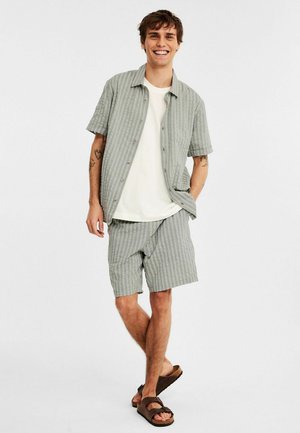 Lys grå kortærmet skjorte med lodrette striber, parret med matchende shorts. Bæret med brune sandaler. Glat stof, afslappet pasform.