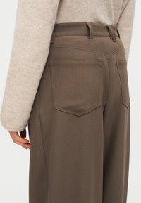 Καφέ ραμμένο παντελόνι με ψηλή μέση, ίσια πόδια και δύο πίσω τσέπες. Το ύφασμα έχει ομαλή, ματ υφή.