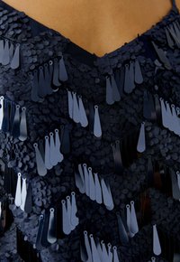 Marineblaues Oberteil mit V-Ausschnitt, das mit geschichteten, ovalen Pailletten und flachen, tropfenförmigen Akzenten verziert ist, was eine strukturierte Oberfläche bietet.