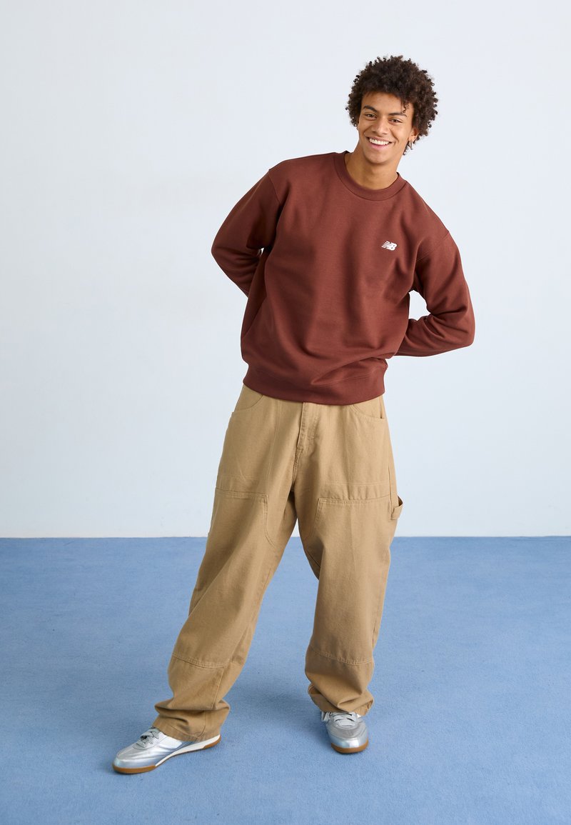 Brauner Pullover mit Logo, kombiniert mit lockeren, beigen Cargo-Hosen. Das Model trägt silberne Sportschuhe und steht auf einem blauen Boden.