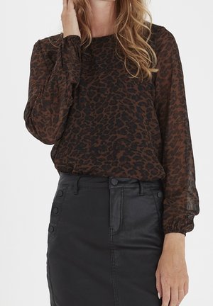 Kobieta ubrana w brązową i czarną przezroczystą bluzkę w panterkę z długimi rękawami oraz wysoką czarną skórzaną spódnicę z guzikami.