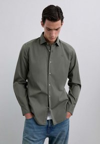 Camisa de botones de manga larga, color verde oliva, hecha de un tejido suave. Cuenta con un cuello, un pequeño logo en el pecho y se combina con jeans de color azul claro.