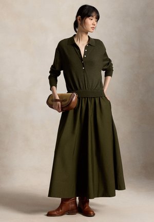 Frau trägt ein dunkelolivfarbenes, langärmeliges, geknöpftes Oberteil und einen passenden langen Rock, hält eine beige-braune Handtasche und trägt braune Stiefeletten.