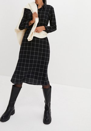 Donna che indossa un vestito midi nero con motivo a griglia bianca, stivali neri al ginocchio e un maglione lavorato a maglia crema drappeggiato sulla spalla.