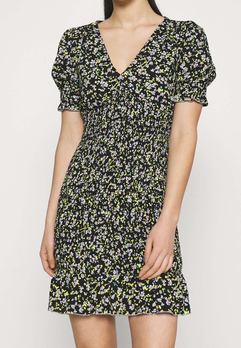Femme portant une robe courte noire à fleurs avec des manches bouffantes, un col en V, une taille smockée et un ourlet à volants.