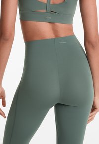 Oliveno zeleni komplet za aktivne aktivnosti, ki vključuje crop top z izrezi in ujemajoče se legice z visokim pasom, izdelane iz gladkega, raztegljivega materiala.