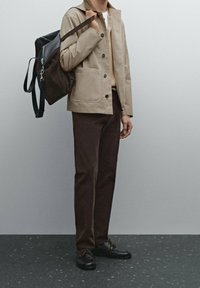 Beige canvas jack met knoopsluiting, crème trui eronder, bruine broek en zwarte schoenen met bruine veters; draagt een donkere rugzak.