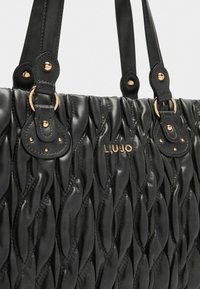 Borsa nera con design trapuntato, caratterizzata da hardware dorato, manici superiori doppi e dettagli di branding. La texture sembra morbida e liscia.