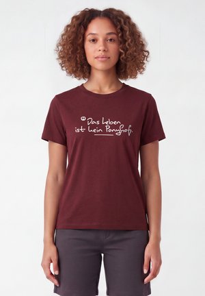 Frau mit lockigem Haar, die ein burgunderfarbenes Shirt mit deutschem Text "Das Leben ist kein Ponyhof" und dunkle Shorts vor einem schlichten weißen Hintergrund trägt.