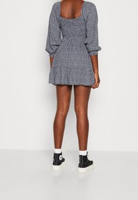 Šaty s květinovým potiskem, čtvercovým výstřihem na zádech, nabíranými rukávy a přiléhavým korpusem, přecházející do rozšířené sukně. Spárováno s černými vysokými sneakers.