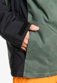 Chaqueta impermeable negra y verde con acabado texturizado, cuenta con una mano en el bolsillo, puños con Velcro y costuras en contraste.