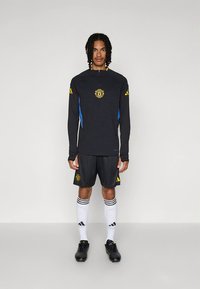 Črna športna majica z dolgimi rokavi in rumeno logotipom, v kombinaciji s črnimi kratkimi hlačami, belimi nogavicami s črnimi črtami in črnimi športnimi čevlji.