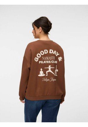 Kvinde iført brun sweatshirt med "Good Day Namaste Pilates Club Tokyo, Japan" og yoga-illustrationer på ryggen.