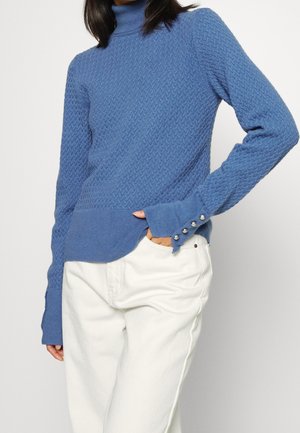 Femme portant un pull à col roulé bleu texturé avec des boutons sur les poignets et un pantalon blanc, debout avec une main dans la poche.