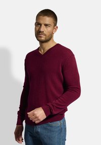 Burgunderfarbener V-Ausschnitt-Pullover aus weichem Material, mit langen Ärmeln und gerippten Bündchen. Kombiniert mit blauen Jeans für einen lässigen Look.