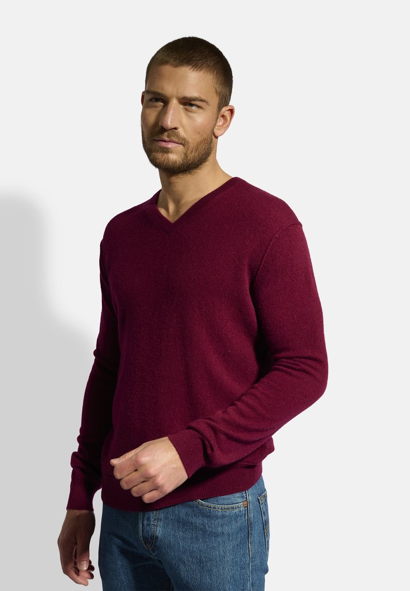 Burgunderfarbener V-Ausschnitt-Pullover aus weichem Material, mit langen Ärmeln und gerippten Bündchen. Kombiniert mit blauen Jeans für einen lässigen Look.
