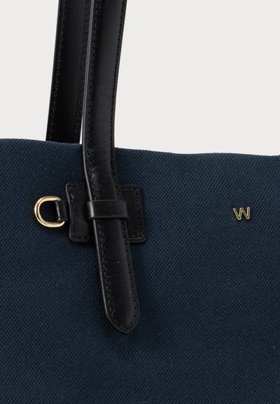 Κοντινή όψη υφασμάτινης τσάντας tote σε ναυτικό μπλε με υφή, με μαύρες δερμάτινες λαβές και μικρό χρυσό λογότυπο "W" στο πάνω δεξιά μέρος.