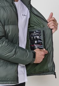 Chaqueta de plumas verde con cremallera, bolsillo interior que muestra una etiqueta negra con iconos y texto. La tela tiene una textura suave y brillante.