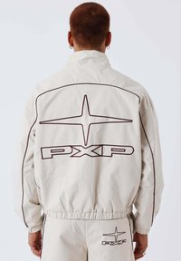 Persoon met kort haar draagt een lichtbeige jas en bijpassende broek, beide met een grote donkere ster met omlijning en het "PXP"-logo op de achterkant.