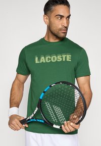 Camiseta verde con la palabra "LACOSTE" impresa en un tono verde más claro, acompañada de una raqueta de tenis negra con detalles en azul y patrón de cuerdas blancas.