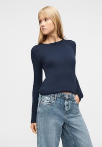 Νεαρή γυναίκα με μπλε σκούρο ριμπ πουλόβερ και ανοιχτόχρωμα τζιν, με το χέρι στην τσέπη, κοιτάζοντας αριστερά της πάνω σε απλό φόντο.