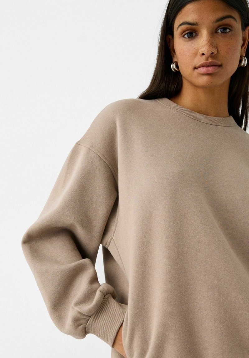 Felpa beige realizzata in morbido tessuto, con scollatura rotonda, spalle cadute e polsini a costine. Nessun motivo o logo visibile.