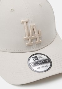 Beige Kappe aus Stoff mit einem gebogenen Schirm. Mit einem strukturierten LA-Logo in passender Farbe und einem verstellbaren Riemen auf der Rückseite.