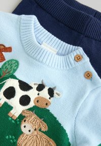 Pull en tricot bleu clair avec des vaches brodées et de l'herbe verte ; boutons en bois au niveau du col ; associé à un pantalon côtelé bleu marine.