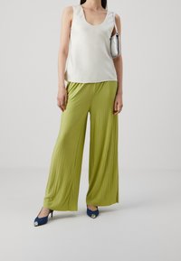 Weißes Satin-Oberteil mit rundem Ausschnitt, kombiniert mit plissierten, weiten limegrünen Hosen. Model trägt marineblaue High Heels mit metallischen Akzenten.