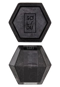 Savon en charbon noir hexagonal avec logo gravé. Emballé dans une étiquette noire comportant du texte et des éléments de design géométriques.