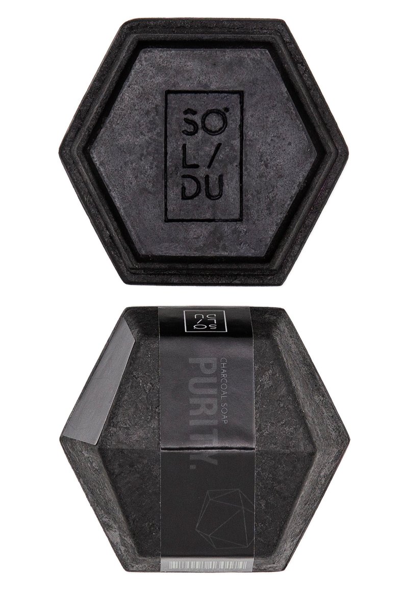 Savon en charbon noir hexagonal avec logo gravé. Emballé dans une étiquette noire comportant du texte et des éléments de design géométriques.