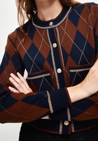 Strickjacke mit geometrischem Muster in Braun, Marineblau und Gold. Mit Rundhalsausschnitt, Frontknöpfen und zwei Brusttaschen.