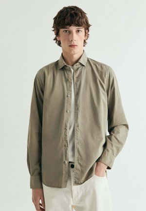 Joven con cabello castaño ondulado lleva una camisa verde oliva abotonada sobre una camiseta blanca, de pie con una mano en el bolsillo de pantalones color crema.