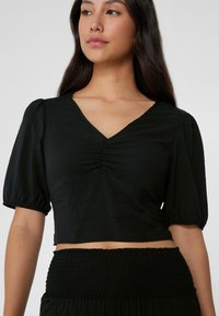 Top negro corto con mangas abullonadas, escote profundo en V y detalle fruncido en la parte delantera. Combinado con una cintura de tela negra fruncida. Textura suave.