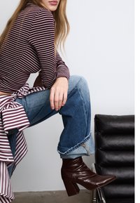 Žena v pruhovaném dlouhém rukávu a zkrácených džínech sedící bokem, oblečená v tmavě hnědých kožených kotníkových botách, poblíž černé kožené židle.