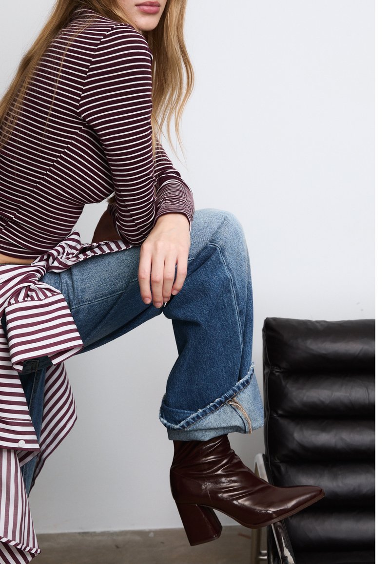 Žena v pruhovaném dlouhém rukávu a zkrácených džínech sedící bokem, oblečená v tmavě hnědých kožených kotníkových botách, poblíž černé kožené židle.