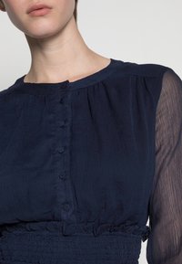 Marinblus med en samlad textur, rund halsringning, knäppning fram och genomskinliga ärmar. Volangdetalj i nederkant.