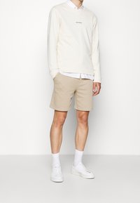 Beige cotton sweatshirt with "LES DEUX" logo, layered over a white button-up shirt. Paired with beige shorts and white sneakers.
