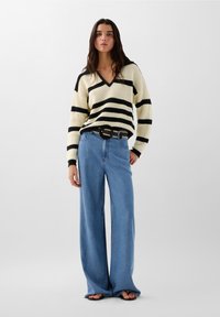 Pull en tricot avec des rayures noires et crème, décolleté en V, associé à un jean bleu taille haute et à jambes larges, et une ceinture noire avec une boucle décorative.