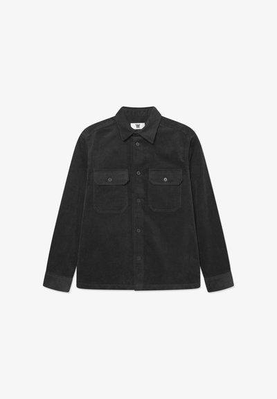 Chemise noire en velours côtelé à manches longues, avec col, fermeture boutonnée sur le devant et deux poches poitrine boutonnées, posée à plat sur fond blanc.
