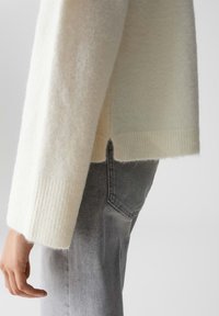 Maglione a maglia crema con polsini a coste, caratterizzato da una vestibilità ampia e fessure laterali. Abbinato a jeans grigi, che presentano una trama liscia e un gradiente di colore.