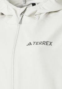 Veste d'extérieur blanche avec fermeture éclair à l'avant, capuche ajustable et un logo "TERREX" gris sur la poitrine gauche. Tissu lisse et léger.