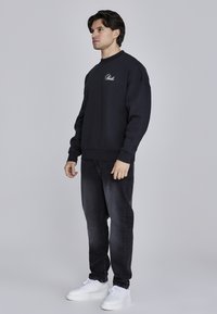 Čierna mikina s rebrovanými manžetami a výstrihom, s bielym vyšitém logom na hrudi, kombinovaná s tmavými džínsami a bielymi teniskami.