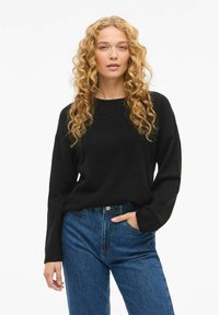 Ung person med långt lockigt blont hår, klädd i en svart tröja och blå jeans, med en hand i fickan mot vit bakgrund.